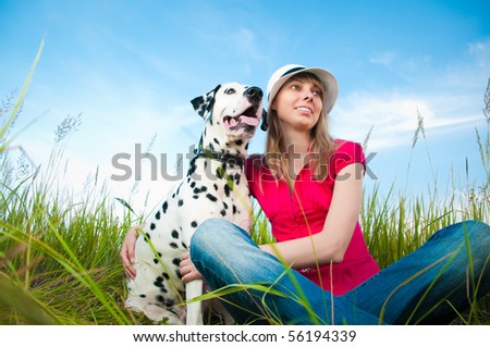 beautiful young woman in hat sitting in grass with her dalmatian dog pet and smiling. Blue sky in background and green grass in foreground.