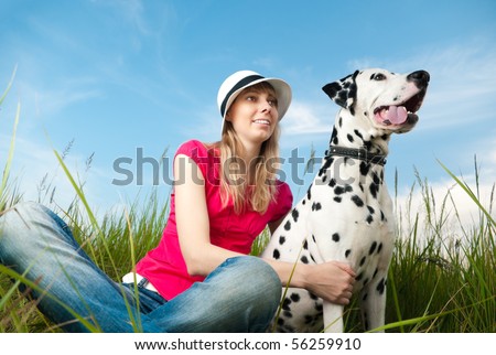 beautiful young woman in hat sitting in grass with her dalmatian dog pet and smiling. Blue sky in background and green grass in foreground.