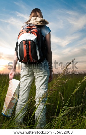 beautiful young woman with backpack and map in hand standing outside in the field with her back to camera. Sunset cloudy blue sky in background and green grass in foreground