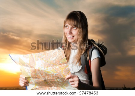 beautiful young woman with backpack and map in hands standing outside in the field and looking in front of her. Sunset cloudy sky in background.