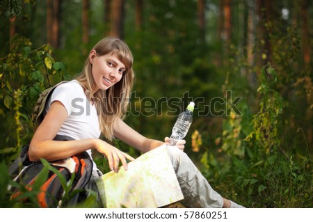 beautiful young woman sitting on  forest floor with backpack, map and bottle of water, smiling and looking in camera
