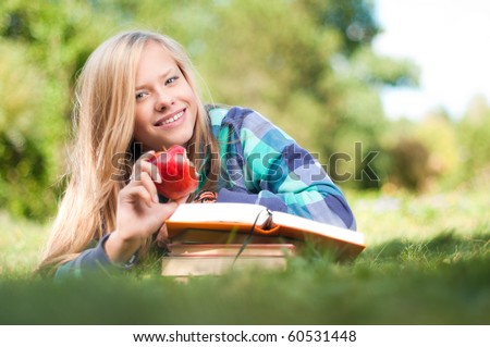beautiful young student girl lying on grass with red apple in her hand and books under her hands, looking into the camera and smiling