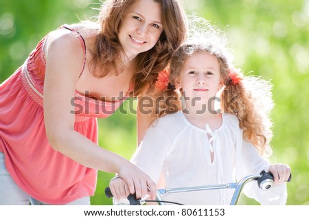 Beautiful and happy young mother teaching her daughter to ride a bicycle. Both smiling and looking into the camera. Summer park in background.