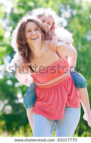Beautiful and happy young mother giving piggyback ride to her daughter. Both smiling. Summer park in background.
