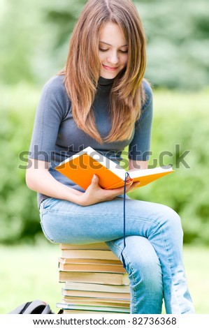 beautiful and happy young student girl sitting on pile of books, holding book in her hands, smiling and reading. Summer or spring green park in background