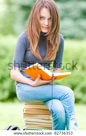 beautiful and happy young student girl sitting on pile of books, holding book in her hands, smiling and looking into the camera. Summer or spring green park in background