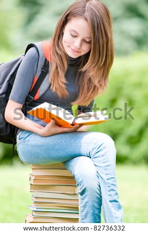 beautiful and happy young student girl sitting on pile of books, holding book in her hands and reading. Backpack on her shoulder. Summer or spring green park in background