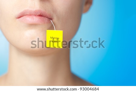 Tea bag in mouth of young woman, yellow label is showing up. Blue background.