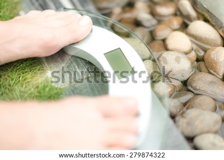 Person on scale with only feet to be seen on green lawn and wet pebble background. Person measures weight in spa setting. Health care and weight loss. Spa helps to be fit.