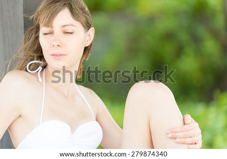 Young sexy woman in white swimsuit relaxing. Pretty girl sitting on beach with her leg pulled up, blurry greenery in background. Summer holidays and vacation. Hotels and tropical resorts.