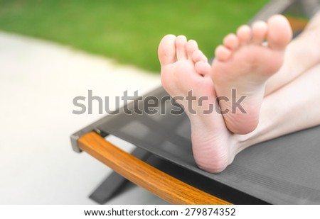 Focus on crossed feet of person lying and tanning on gray sunbed. Green lawn in background. Woman\'s legs with smooth skin. Person relaxing and sunbathing. Leisure activity, vacation.