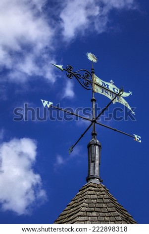 Weather vane on a sunny blue sky with white fluffy clouds. Wind direction arrow points west and has a slight green patina on the copper finial