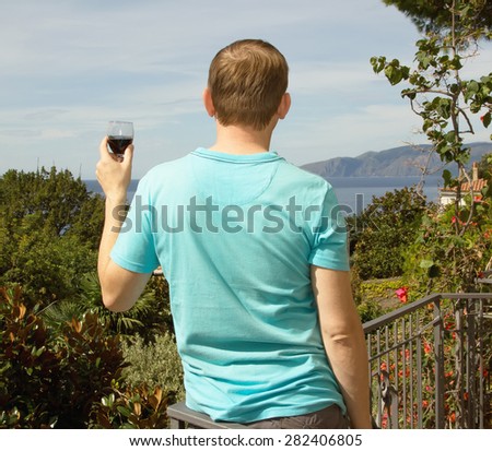 Man drinking red wine from glass, he is on vacation. The man against the sky, the sea, mountains and green plants. Man tasting wine. View from the back. It stands - rear view. South. Europe.