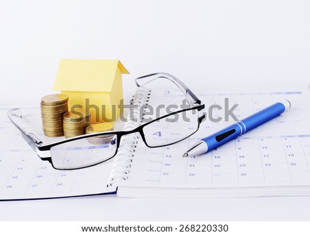 Loans concept with Eyeglasses and blue pen and money coins and yellow paper house on calendar book page
