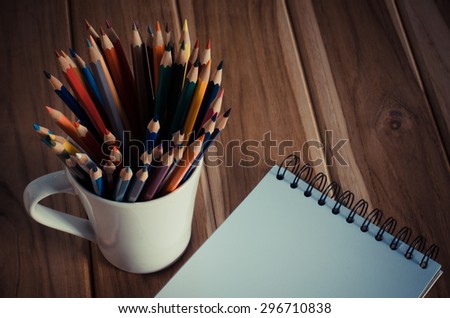 Color pencils  in the cups and notebook placed on a desk.