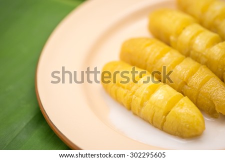 Fresh and delicious boil yellow banana with coconut milk on brown metal plate on banana green leaf for local Thai dessert background