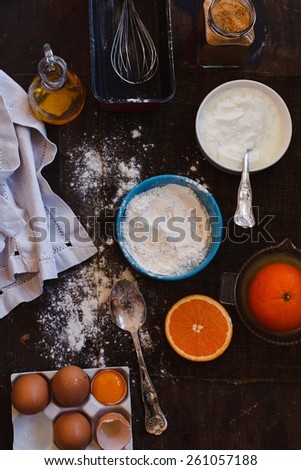 Assorted baking ingredients and tools:  flour in measuring cup, eggshels, cup of yogurt, juicer of orange, taken over dark wood table. Homemade Baked biscuit cake. Step on step.  Top view.