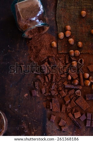 A stack of different chocolates, lots of grated chocolate, slices of dark and milk chocolate, cacao powder and hazelnut kernels, taken on dark vintage table. Rustic style. Top view.