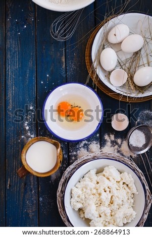 Homemade fresh ricotta cottage cheese made from milk with eggs and jug of milk over on blue wooden table. Rustic style