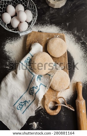 Three balls of dough over vintage cutting board and linen napkin from above dark board with bowl of eggs and rolling pin. Rustic composition style.