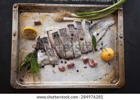 Cooked fish concept. Preparing white fillet fish with lemon, green garlic, olive oil and seasonal spice on kitchen tray over dark background. Top view.