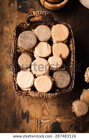 Many jars of canned goods located in rustic woven willow basket from above over on vintage dark table. Rustic atmosphere from natural sunlight.