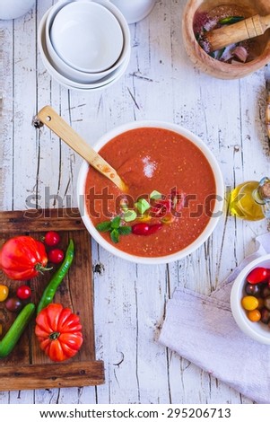 A Delicious cold gazpacho soup in white bowl with bowl tomatoes cherry and bottle of olive oil over on a rustic white wooden kitchen table from above. Traditional spanish cold soup. Step on step.