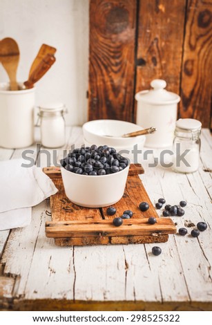 A white bowl of fresh harvest blueberry over on vintage chalkboard from above on white rustic kitchen table with white ceramic cookware and spoon. Rustic styling.