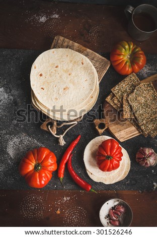Stack of homemade whole flour tortilla with tomatoes and crust tortilla on vintage cutting board with chili red pepper, from above on dark stone table. Rustic dark style