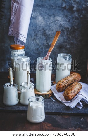 Real country dairy food concept. Preparing homemade kefir and natural yogurt served in glass jars over stone blue table with whole grain bread over on rustic linen cloth. Dark rustic styling scene.