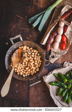 A pan with cooked chickpeas and grilling various raw sausages from above on rustic ages table with small peppers . Rustic dark styling. Country typical homemade food.