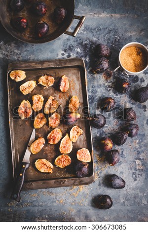 A Succulent figs scattered above cooking sheet form above on rustic aged table with bowl of sugar. Rustic dark styling.