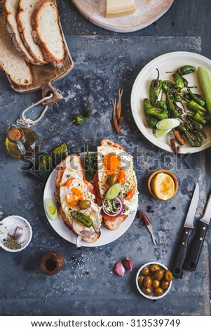 A Toasted sandwich with smoked salmon and fried eggs and variety cheese cutting board with bowl olives and green leaves salad from above a blue rustic table. Country simple mediterranean food .