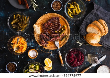 Succulent thick juicy portions of grilled fillet steak served with variety veggies dips, fermented veggies, cucumber and pepper marinated, bread buns on a old vintage table  Authentic dinner party.