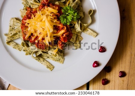 Delicious green herb pasta with bolognese sauce on white plate with red small heart-shaped pebbles, wooden background, top view