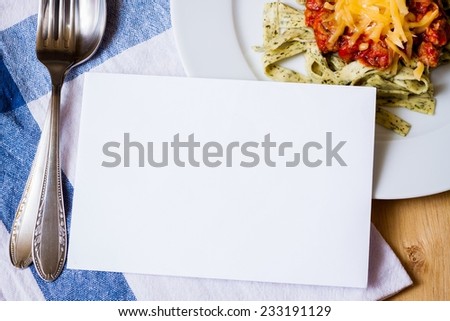 Blank card with delicious green herb pasta and bolognese sauce on white plate with fork, spoon and dishtowel
