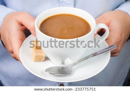 Hand holds mug of coffee with milk with spoon and brown sugar