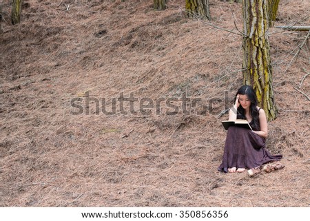 Young woman in long dress reads a book sitting under a tree