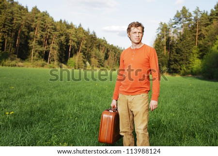 Handsome young man with a suitcase standing in the sunshine in a green grassy field with pine trees in the distance