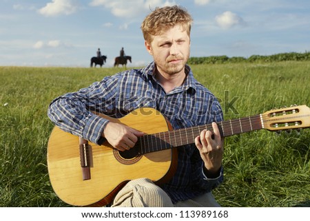 Young blonde man strumming a guitar while sitting in an open grassy field with horse riders on the skyline