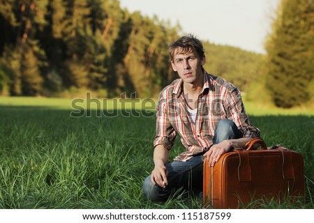Man sitting with his suitcase in grass Casual young man kneeling alongside his suitcase in a green grass field in evening light