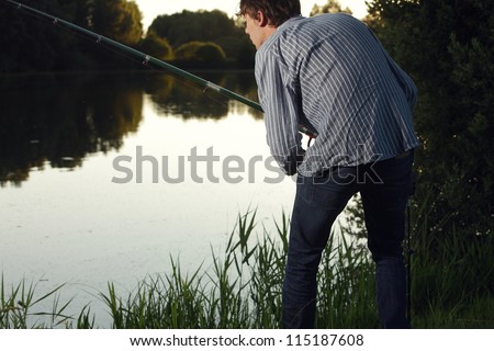 Man fishing at lake at sunset Young man standing on a grassy bank fishing at a lake in evening light
