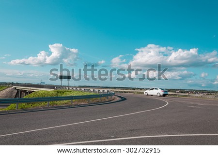 the white car at a speed passes a road fork against the blue sky and fluffy clouds