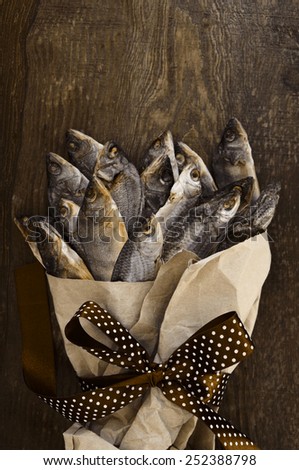 Salty dry river fish is wrapped in crumpled yellow paper and decorated with a brown tape with a bow on a brown wooden background / ?he Bouquet from salty dry fish. A gift for men.