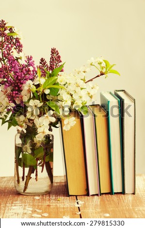 Pile of books and spring flowers of cherry and lilac in a glass jar on a light background on a wooden surface. Composition with books and spring flowers