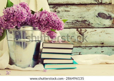 Bouquet of lilac flowers in a metal bucket and a pile of books against an old wooden board, a decor in vintage style