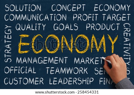 Economy word cloud is written on blue chalkboard by the hand of a businessman.