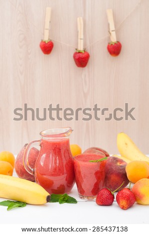 Healthy strawberry banana smoothie with mint in a glass and pitcher on wooden background. Fresh fruits bananas, peaches and apricots background. Selective focus