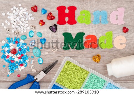 Crystals, pendants, charms, plier, glass hearts, box with beads, accessories to create hand made jewelry and word handmade of felt on wooden background. Tools for creating jewelry