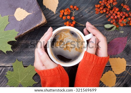 Female hands holding cup of coffee on autumn wooden background. Old book, autumn leaves and dried rowan on background. Concept cozy coffee cup. Top view